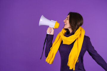 Young woman in a knitted dress and in a yellow scarf on a purple background cheerfully shouts into a megaphone, cheerful, announces a sale