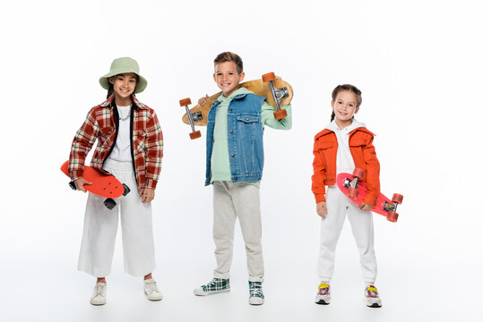 full length of trendy children smiling while holding longboard and penny boards on white.