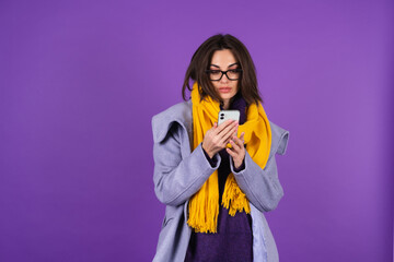 Young woman in a gray coat, knitted dress and in a yellow scarf on a purple background, spring and winter style, fashionable glasses, looks at the phone screen