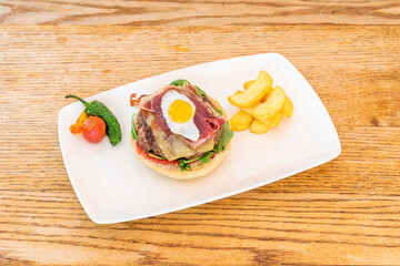 Beef burger with melted cheese, slices of Serrano ham, arugula, quail egg on top and fried peppers with potatoes