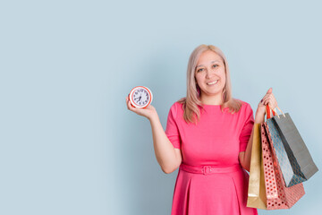 An adult 40-year-old woman with an alarm clock and shopping bags stands on a blue background,...