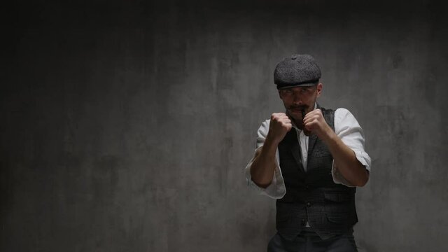 Young dangerous gangster man dressed in vintage suit, cap and smoking pipe, rolls up his sleeves, prepares for fight, waving his fists, provoking, stands in fighting stance, in front of concrete wall
