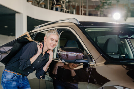 Happy smiling woman customer female buyer client in black shirt looking in mirror car choose auto want buy automobile in car showroom vehicle salon dealership store motor show indoor. Sales concept.