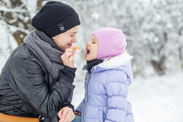 Mom gives her little daughter a tangerine. Walk in the snowy park.
