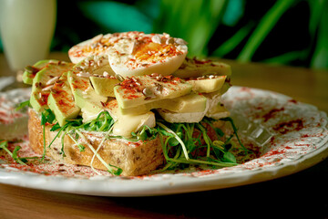 Avocado toast with brie cheeese, rucola and poached egg in white plate on wooden background