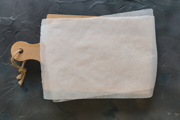 Wooden cutting board and white parchment paper on dark gray background. Mockup for recipes,...