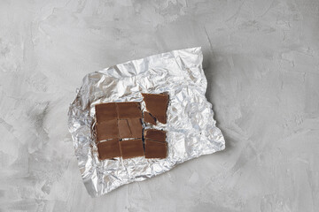 Pieces of broken milk chocolate bar on silver foil wrapper, gray background. Ingredient for cooking. Top view, copy space