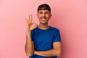 Young mixed race man isolated on pink background winks an eye and holds an okay gesture with hand.