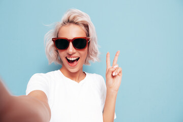 Young beautiful smiling female in trendy summer clothes. Sexy carefree woman posing near blue wall in studio. Positive blond model having fun in sunglasses. Cheerful and happy. Taking Pov selfie