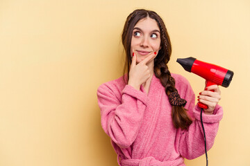 Young woman holding a hairdryer isolated on yellow background looking sideways with doubtful and skeptical expression.