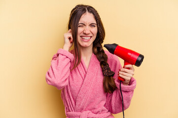 Young woman holding a hairdryer isolated on yellow background covering ears with hands.