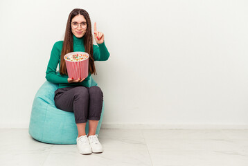 Young caucasian woman eating popcorns on a puff isolated on white background showing number one with finger.