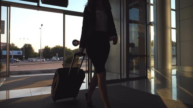 Business lady going to the airport with her luggage. Young woman in heels entering walking through glass doors to the terminal and roll suitcase on wheels. Trip or vacation travel concept. Slow motion