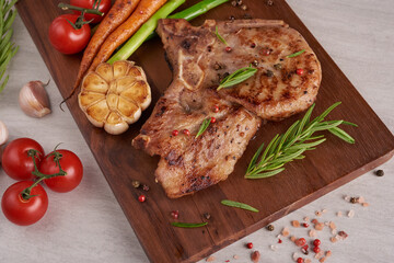 Grilled pork steak from a summer BBQ served with Vegetables, asparagus, baby carrots, fresh tomatoes and  spices. Grilled steak on wooden cutting board  on stone background . top view.