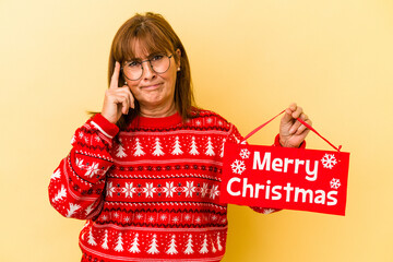 Middle age caucasian woman holding ‘’merry Christmas’’ placard isolated on yellow background pointing temple with finger, thinking, focused on a task.