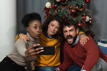 Three young friends celebrate Christmas and New Year's Eve Holidays together by joking and taking selfies with a smartphone goofy and making funny facial expressions in front of the Christmas tree.  - obrazy, fototapety, plakaty