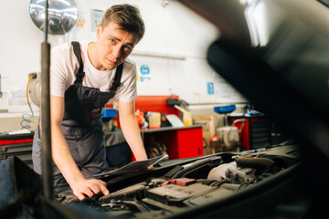 Portrait of confident handsome professional male car mechanic in blue uniform standing in front of open hood, inspecting engine of car coming in for repair or maintenance in auto repair workshop..