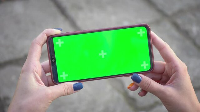 Woman holds mobile phone and clicks on screen swipes photos or pictures on street on background of paving slabs. Chroma key mock-up on smartphone in hand. Use green screen for copy space closeup 4k