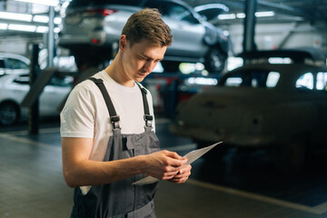 Side view of focused handsome young mechanic male wearing uniform reading clipboard standing in...