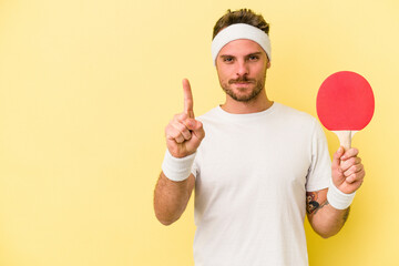 Young caucasian man holding ping pong racket isolated on yellow background showing number one with...