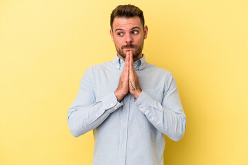 Young caucasian man isolated on yellow background praying, showing devotion, religious person looking for divine inspiration.