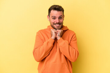 Young caucasian man isolated on yellow background keeps hands under chin, is looking happily aside.
