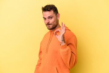 Young caucasian man isolated on yellow background winks an eye and holds an okay gesture with hand.