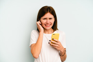 Young English woman holding mobile phone isolated on blue background covering ears with hands.