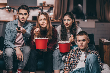 A company of cute young two guys and two girls sitting on the couch, watching TV, eating popcorn, bored, changing the channel