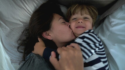 Toddler hugging mother lying in bed together, family love and affection