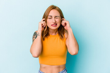 Young caucasian woman isolated on blue background covering ears with hands.