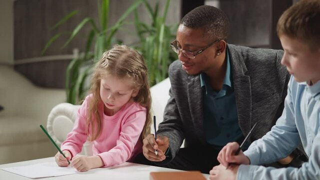Attentive black man teacher sits with little children students doing homework at private English tuition with native speaker at home slow motion