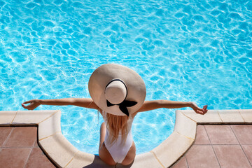 Fototapeta na wymiar Top view of a woman with a sunhat sitting at the pool edge and enjoying the hot summer day