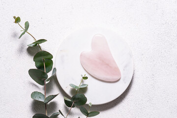 Rose quartz gua sha massager on white marble background with fresh aromatic eucalyptus branches. Skin care concept.