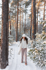 Happy woman walks in beige jacket parka a winter forest with the morning light streaming through the trees and illuminating the pine trees behind.