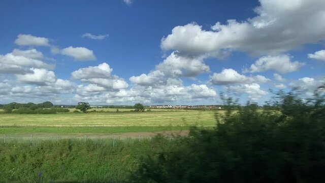 Wide shot out of a window of train, tracking along flat british UK southern England countryside fields and farmland clouds and blue sky - stock footage video