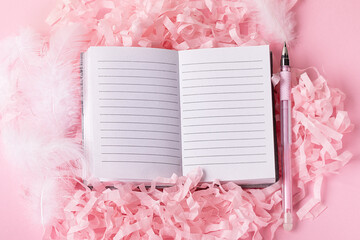 cute pink fluffy background with open notepad and  pen on decorative paper next to it. Top view of on girly stationery and white decorative feathers with ribbons
