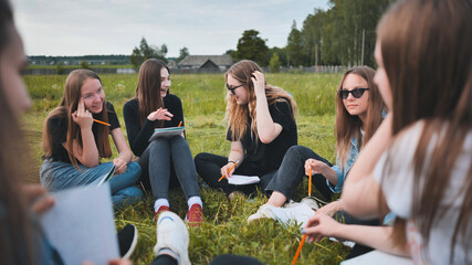 A group of female students are sitting in a circle on a meadow for collective work with notebooks.