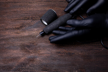 the hands of the permanent makeup artist in black sterile gloves move the tattoo machine with their fingers, which lies on a wooden table