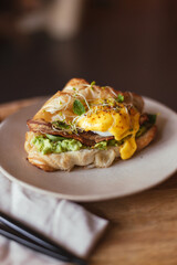 Egg benedict with bacon, micro green sprouts, mashed avocado in croissant. American drip black coffee. Coffee table. Yellow, green colors. Fancy breakfast or brunch. Moody food photography. Fresh food
