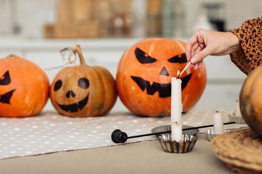 holidays and leisure concept - woman's hand with matches lighting candles at home on halloween. painted pumpkins on a background. horror theme and Hallowe'en. selective focus.