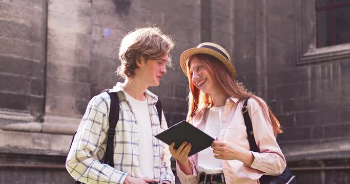 Beautiful female girl holding digital tablet and showing pictures to male friend. Young redheaded woman in hat and guy with backpack looking through photos and laughing. Memories, technology.