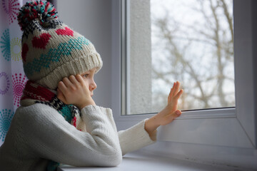 A sad 7-year-old boy in a cap and a scarf is sitting by the window at home.