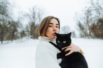 Portrait of stylish woman in white sweater outdoors in winter with black cat in hands posing at camera, focused looking at camera.