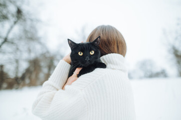 Back of a woman in a white knitted sweater with a black cat on her shoulders on a winter walk....