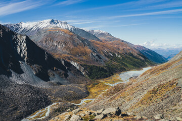 Picturesque highland landscape with mountain river and lake in valley among high mountains with snow in autumn colors in sunshine. Awesome alpine view to motley mountain valley in sunlight in fall.