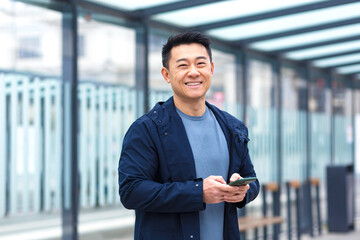 Cheerful and happy asian using phone smiling and looking at camera, near airport, businessman on tourist trip