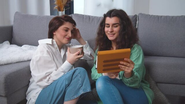 Happy young lesbian couple sitting on home floor, using tablet computer, browsing, laughing at funny photos, videos, having fun together, enjoying their weekend at home. Caucasian women