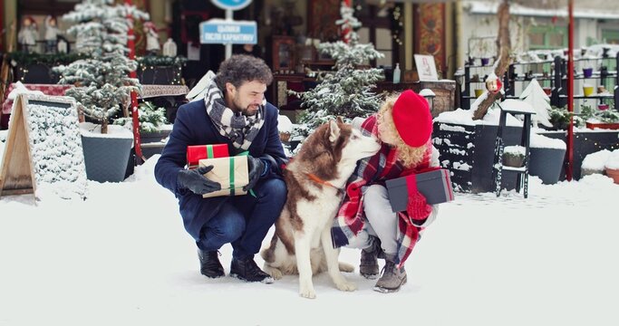 Beautiful young couple sitting with a dog on decorated street, smiling and holding Christmas gifts. Christmas mood concept.