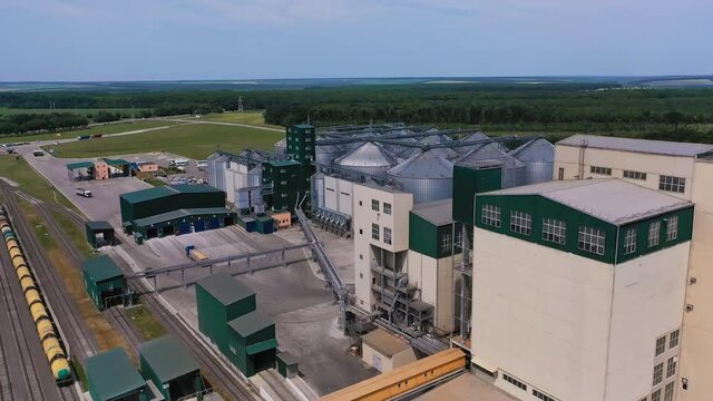 Commercial grain silos outdoors. Flight from the industrial building of the plant to the large metal tanks of elevator. Aerial view. Woods and farmlands at the backdrop.